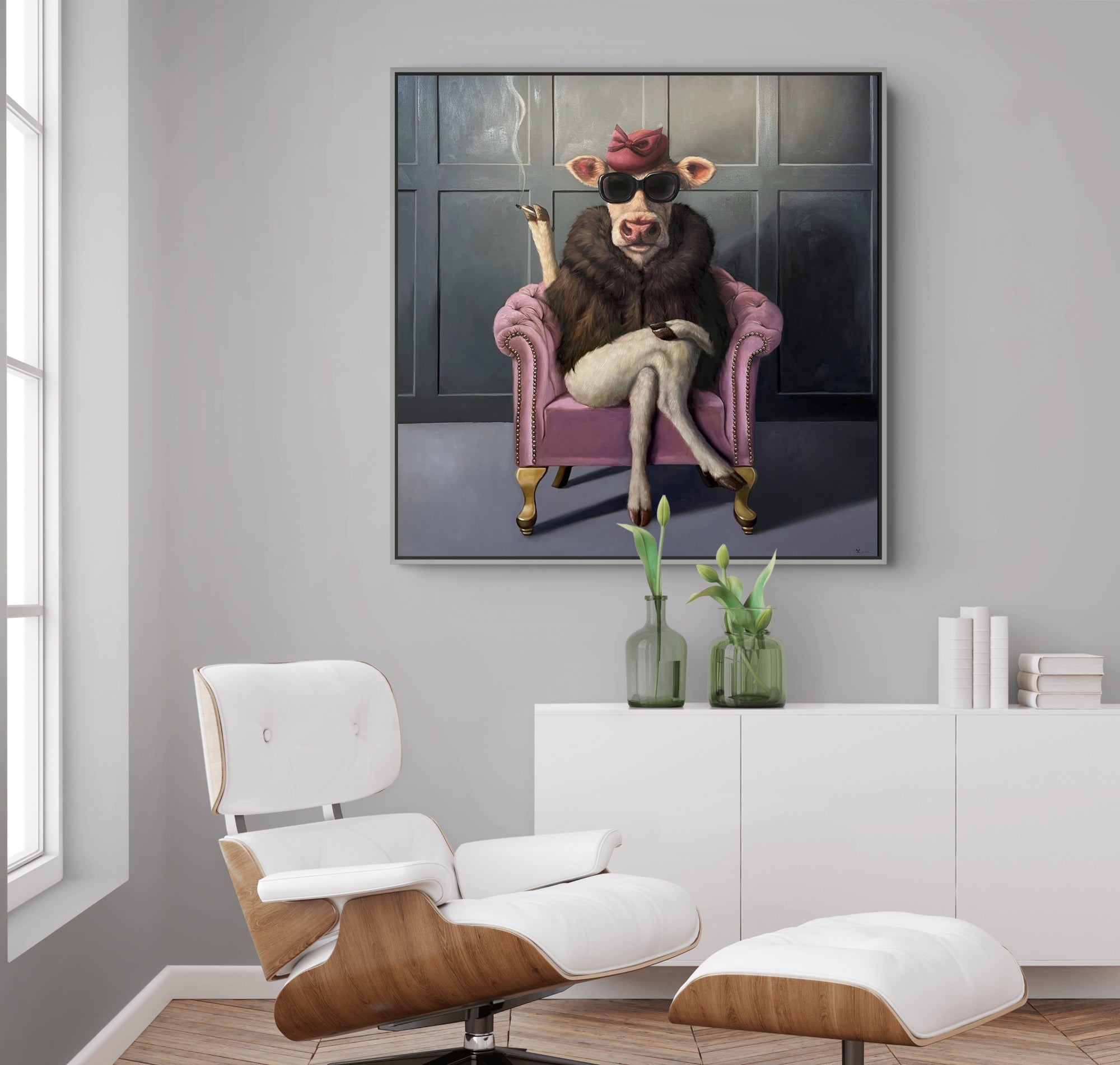 A framed wall art print showing a stylized animal sitting in a pink armchair, displayed above a white sideboard in a modern living room with a white lounge chair and ottoman.